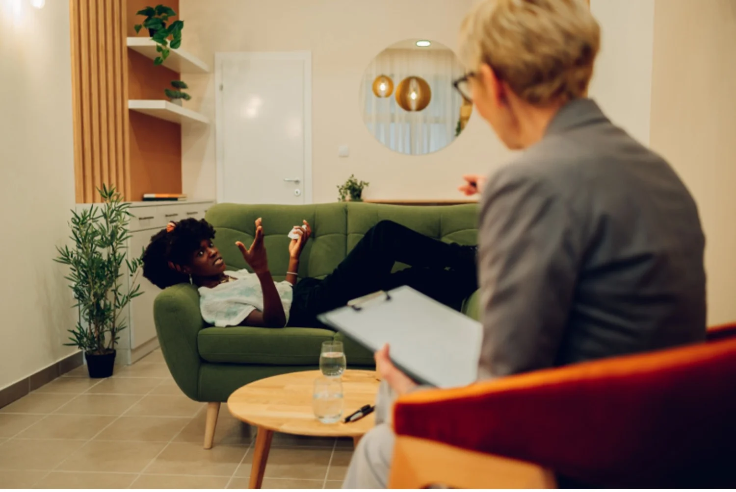 Young woman laying on therapist’s couch gestures with her hands while therapist takes notes